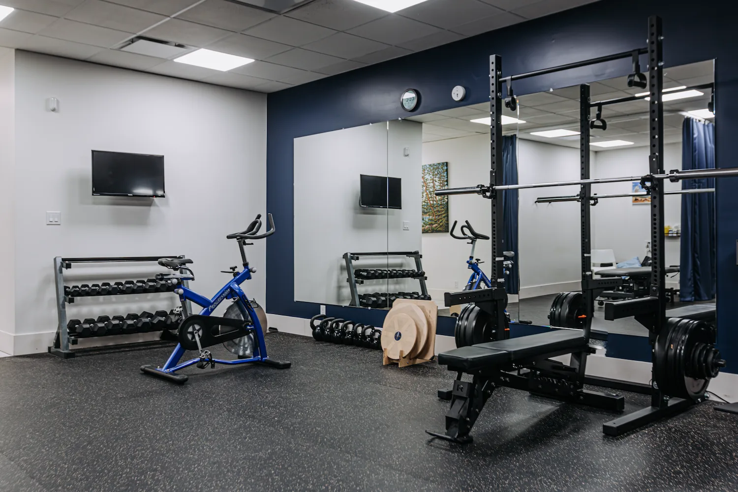 Gym floor at Launch Rehab North Burnaby with squat rack, dumbbell rack, bench, and spin bike against a navy accent wall