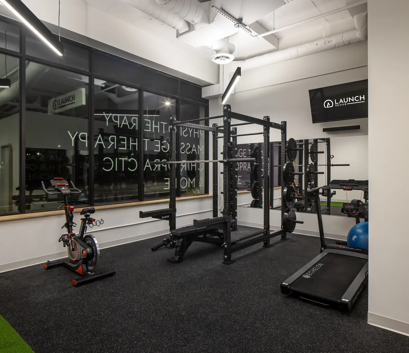 Gym floor at Launch Rehab Richmond with Echelon spin bike, Rogue power squat rack and bench, weight tree, mirror wall, treadmill, and the storefront windows showing the reverse-vinyl Physiotherapy · Massage · Chiropractic & More text