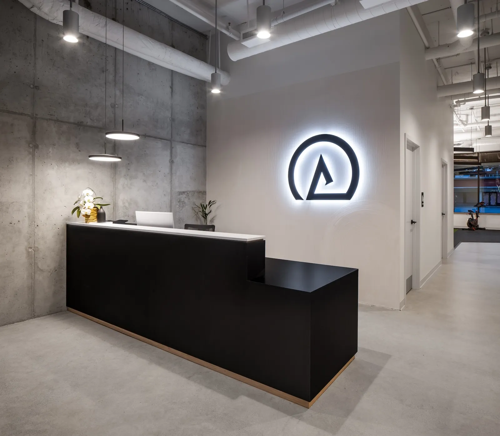 Reception desk at Launch Rehab Richmond: a black L-shaped reception desk with the backlit Λ logo on the white wall behind it, concrete column wall on the left with pendant lights, and the spin bike visible through the doorway to the gym