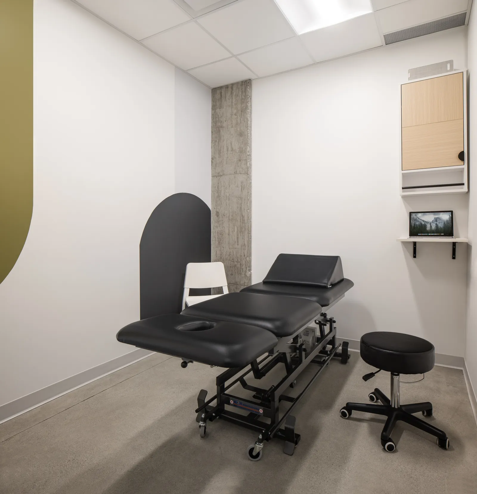 Treatment room at Launch Rehab Richmond with a black KOR Innovations treatment table, white chair, rolling stool, olive accent wall and exposed concrete column, and wall-mounted laptop shelf